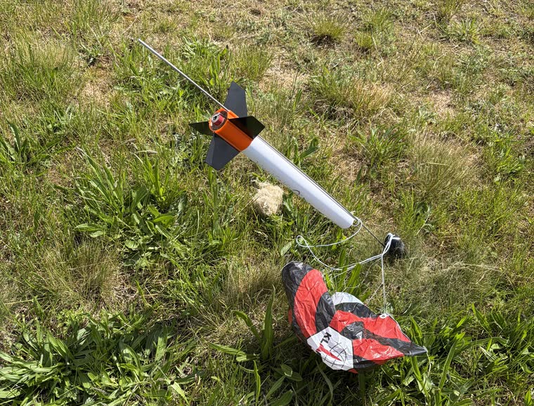Think Celestial lying in the grass beside its red and black parachute, wadding and shock cord still attached, with the launch rod it pulled off the pad still through its lug, 3 minutes after the launch.